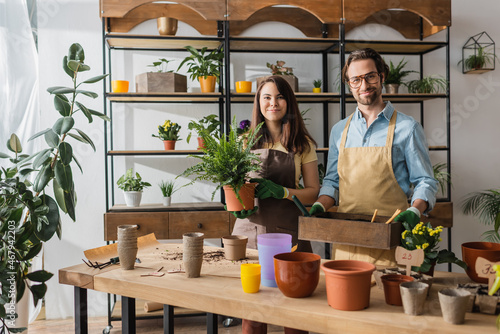 Smiling florists in aprons holding box with ground and plant in flower shop