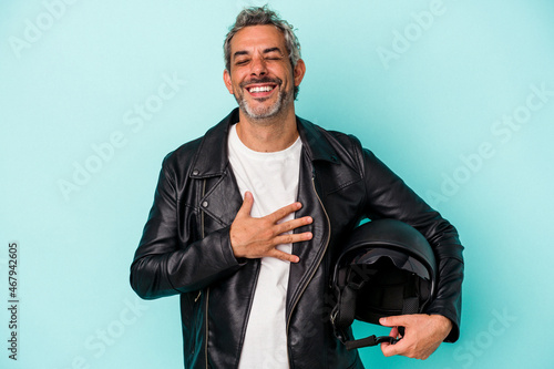 Middle age biker caucasian man holding helmet isolated on blue background  laughs out loudly keeping hand on chest.