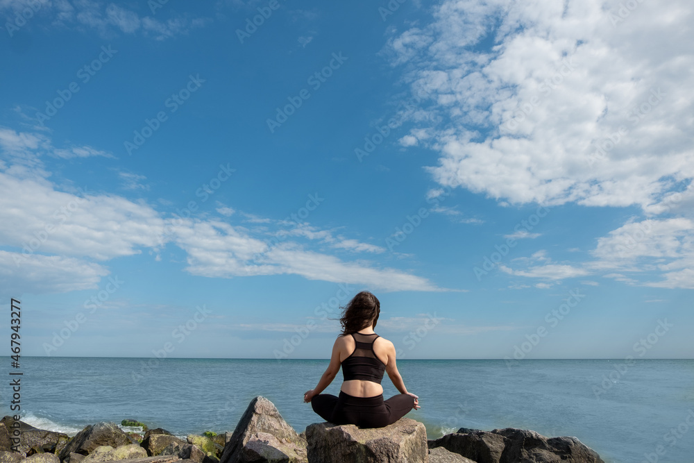 Meditating woman. Peaceful harmony. Yoga practice. Unrecognizable lady sitting in lotus pose on rock stone looking azure water coast blue sky.