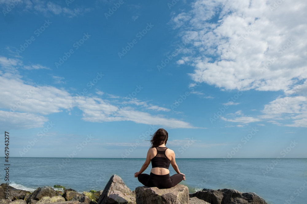 Meditating woman. Peaceful harmony. Yoga practice. Unrecognizable lady sitting in lotus pose on rock stone looking azure water coast blue sky. Meditating woman. Peaceful harmony. Yoga practice. Unrecognizable lady sitting in lotus pose on rock stone looking azure water coast blue sky.