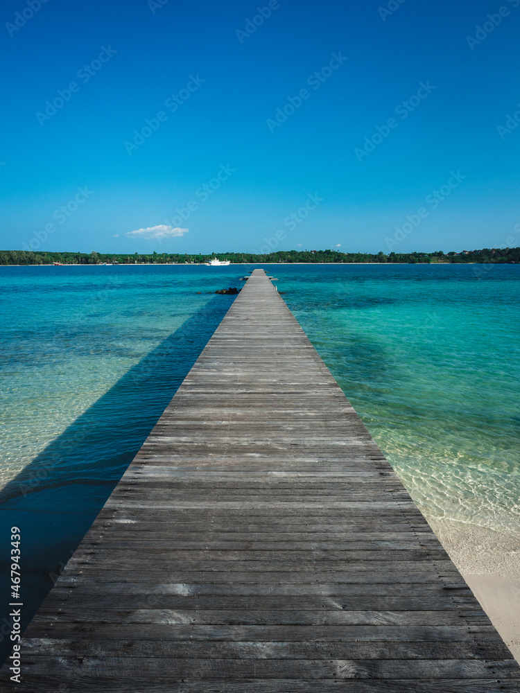 Long straight wooden pier over crystal clear turquoise water and white sand beach of Koh Kham Island. Look to Koh Mak Island, Trat Province, Thailand.