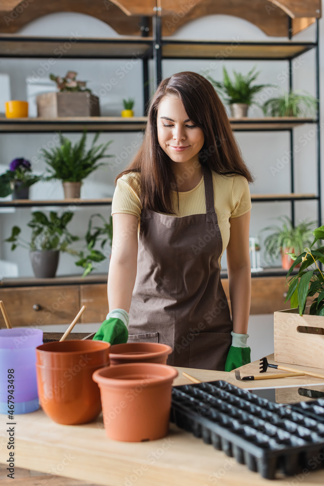 Young florist in gloves holding flowerpots near gardening tools in flower shop