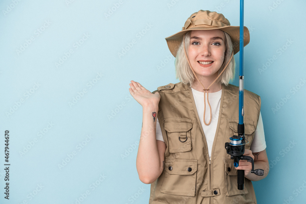 Young caucasian fisherwoman holding a rod isolated on blue background showing a copy space on a palm and holding another hand on waist. Young caucasian fisherwoman holding a rod isolated on blue background showing a copy space on a palm and holding another hand on waist.