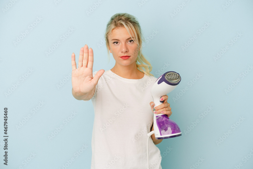 Young Russian woman holding vertical iron isolated on blue background standing with outstretched hand showing stop sign, preventing you. Young Russian woman holding vertical iron isolated on blue background standing with outstretched hand showing stop sign, preventing you.
