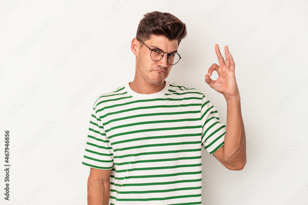 Young caucasian man isolated on white background winks an eye and holds an okay gesture with hand. Young caucasian man isolated on white background winks an eye and holds an okay gesture with hand.