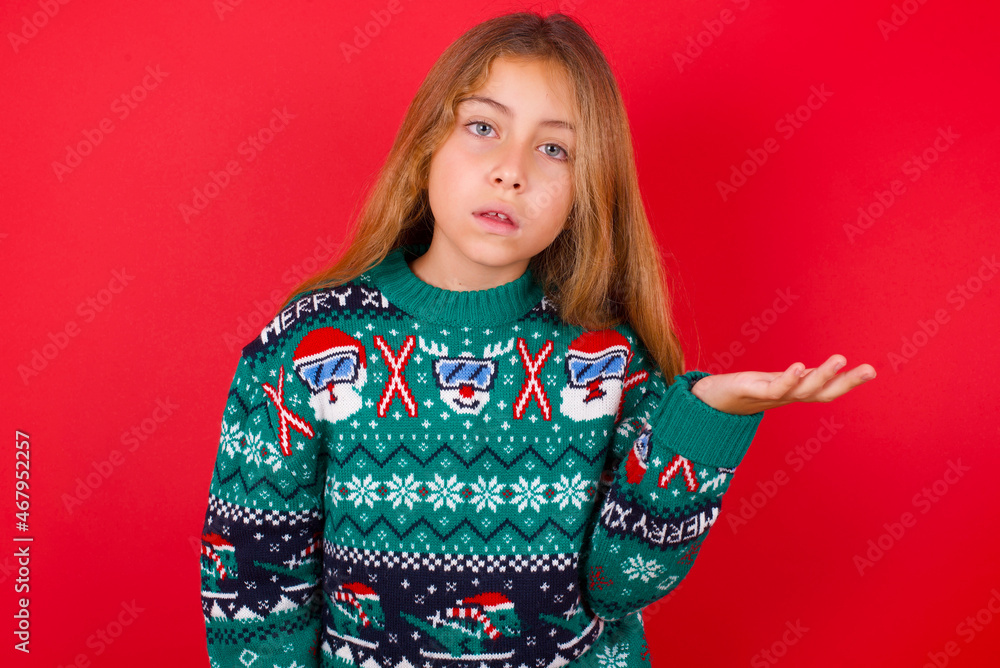 Studio shot of frustrated brunette kid girl in knitted sweater Christmas over red background gesturing with raised palm, frowning, being displeased and confused with dumb question. Studio shot of frustrated brunette kid girl in knitted sweater Christmas over red background gesturing with raised palm, frowning, being displeased and confused with dumb question.