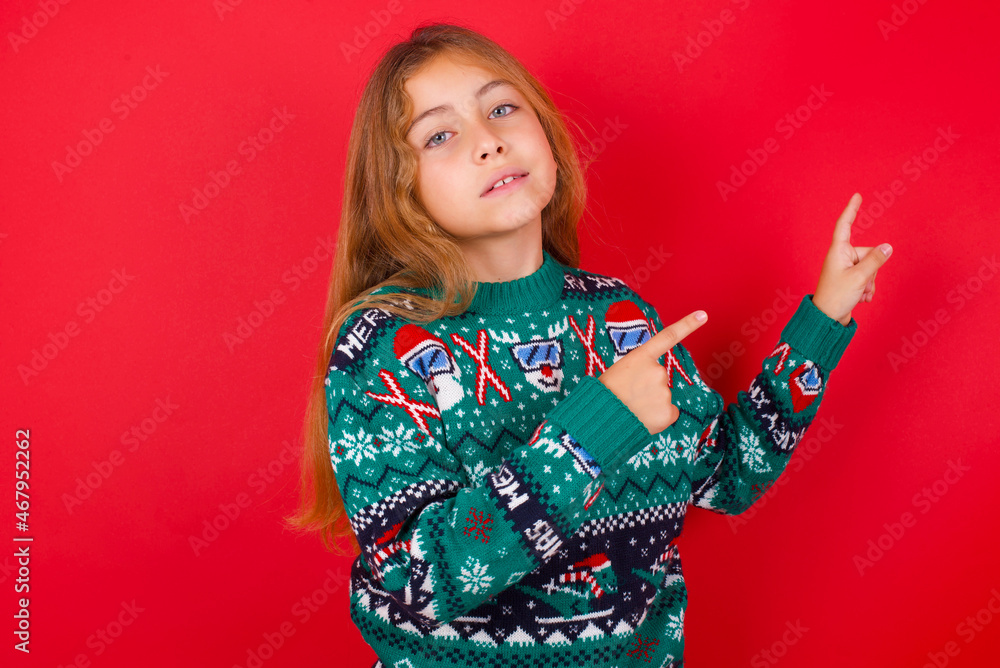 Positive brunette kid girl in knitted sweater Christmas over red background with beaming smile pointing with two fingers and looking on empty copy space. Advertisement concept. Positive brunette kid girl in knitted sweater Christmas over red background with beaming smile pointing with two fingers and looking on empty copy space. Advertisement concept.