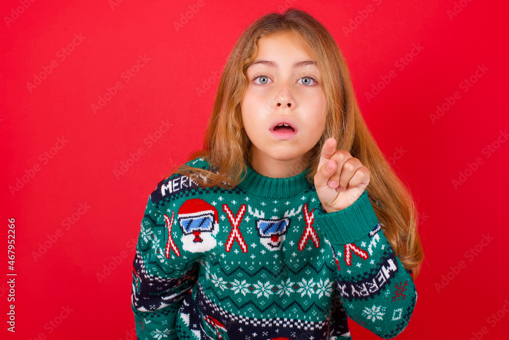 Shocked brunette kid girl in knitted sweater Christmas over red background points front with index finger at camera and. Surprise and advertisement concept. Shocked brunette kid girl in knitted sweater Christmas over red background points front with index finger at camera and. Surprise and advertisement concept.