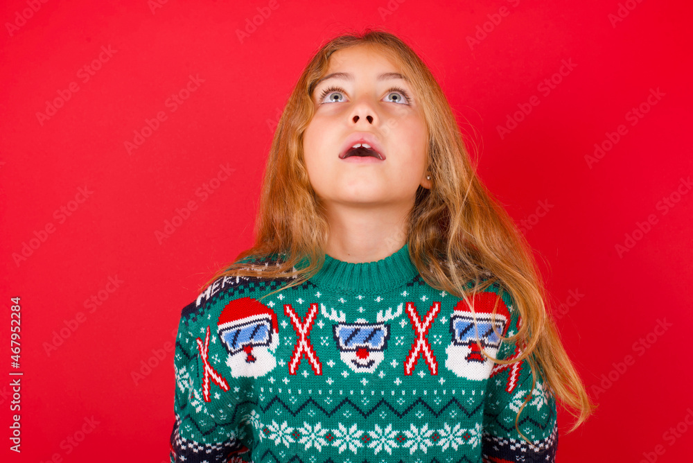 Surprised brunette kid girl in knitted sweater Christmas over red background , shrugs shoulders, looking sideways, being happy and excited. Sudden reactions concept. Surprised brunette kid girl in knitted sweater Christmas over red background , shrugs shoulders, looking sideways, being happy and excited. Sudden reactions concept.