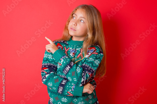 Portrait of brunette kid girl in knitted sweater christmas over red background posing on camera with tricky look, presenting product with index finger. Advertisement concept.
