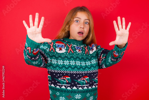 Quadro em tela Dissatisfied brunette kid girl in knitted sweater christmas over red background frowns face, has disgusting expression, shows tongue, expresses non compliance, irritated with somebody