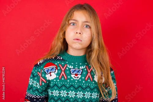 brunette kid girl in knitted sweater Christmas over red background  expressing disgust, unwillingness, disregard having tensive look frowning face, looking indignant with something.