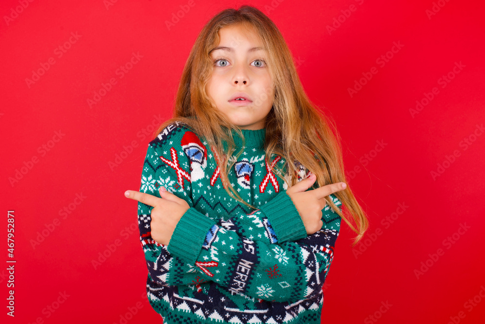 Confused brunette kid girl in knitted sweater Christmas over red background chooses between two ways, points at both sides with crossed hands, feels doubt. Need your advice. Confused brunette kid girl in knitted sweater Christmas over red background chooses between two ways, points at both sides with crossed hands, feels doubt. Need your advice.