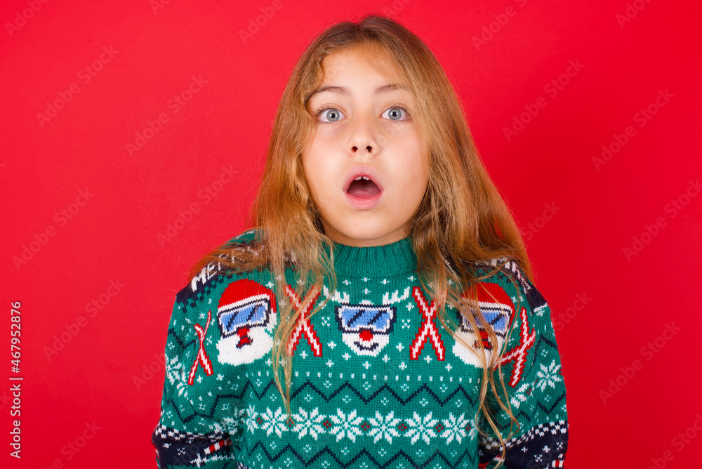 Emotional attractive brunette kid girl in knitted sweater Christmas over red wall with opened mouth expresses great surprise and fright, stares at camera. Unexpected shocking news and human reaction. Emotional attractive brunette kid girl in knitted sweater Christmas over red wall with opened mouth expresses great surprise and fright, stares at camera. Unexpected shocking news and human reaction.