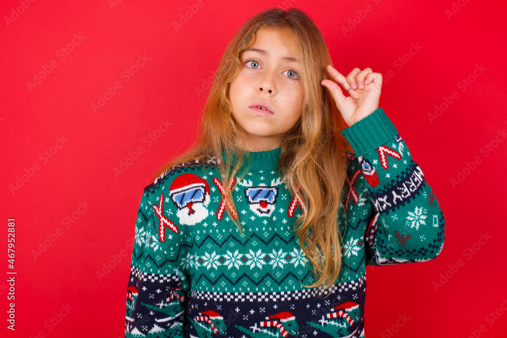 Shocked brunette kid girl in knitted sweater Christmas over red background shows something little with hands, demonstrates size, opens mouth from surprise. Measurement concept. Shocked brunette kid girl in knitted sweater Christmas over red background shows something little with hands, demonstrates size, opens mouth from surprise. Measurement concept.