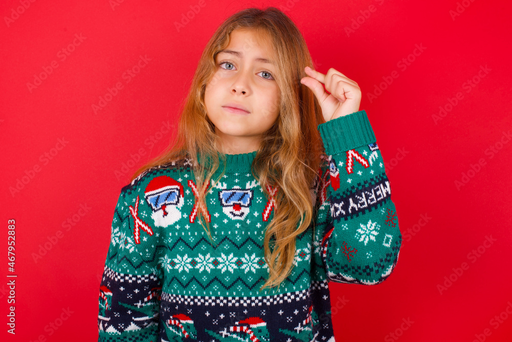 Upset brunette kid girl in knitted sweater Christmas over red background shapes little gesture with hand demonstrates something very tiny small size. Not very much Upset brunette kid girl in knitted sweater Christmas over red background shapes little gesture with hand demonstrates something very tiny small size. Not very much