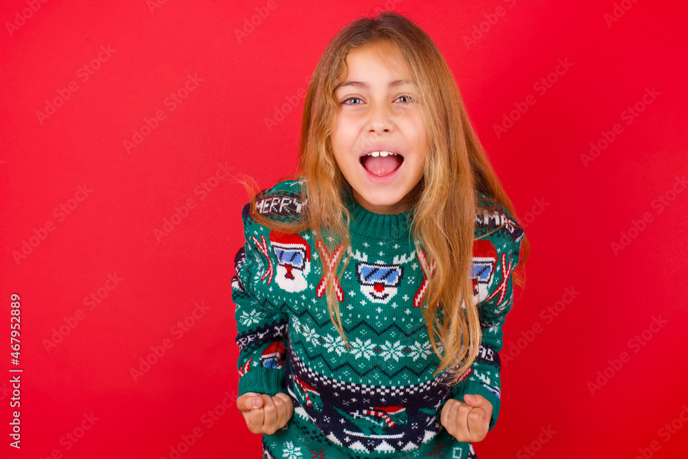brunette kid girl in knitted sweater Christmas over red background raising fists up screaming with joy being happy to achieve goals. brunette kid girl in knitted sweater Christmas over red background raising fists up screaming with joy being happy to achieve goals.