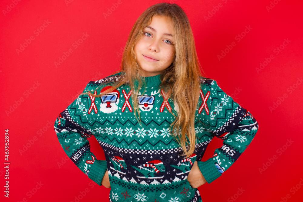 Funny frustrated brunette kid girl in knitted sweater Christmas over red background holding hands on waist and silly looking at awkward situation. Funny frustrated brunette kid girl in knitted sweater Christmas over red background holding hands on waist and silly looking at awkward situation.
