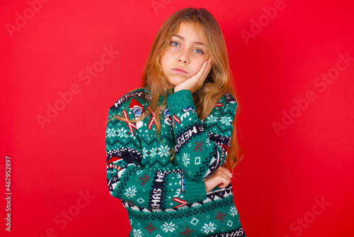 Very bored brunette kid girl in knitted sweater christmas over red background holding hand on cheek while support it with another crossed hand, looking tired and sick.