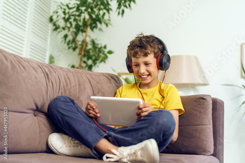 happy boy with headphones and a tablet is sitting at home on couch. child is happy to learn at home online on computer tablet, listening to music. new children's learning app. Dependence on gadgets