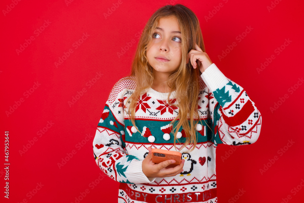 brunette kid girl in knitted sweater christmas over red background holding gadget while sticking out tongue brunette kid girl in knitted sweater christmas over red background holding gadget while sticking out tongue