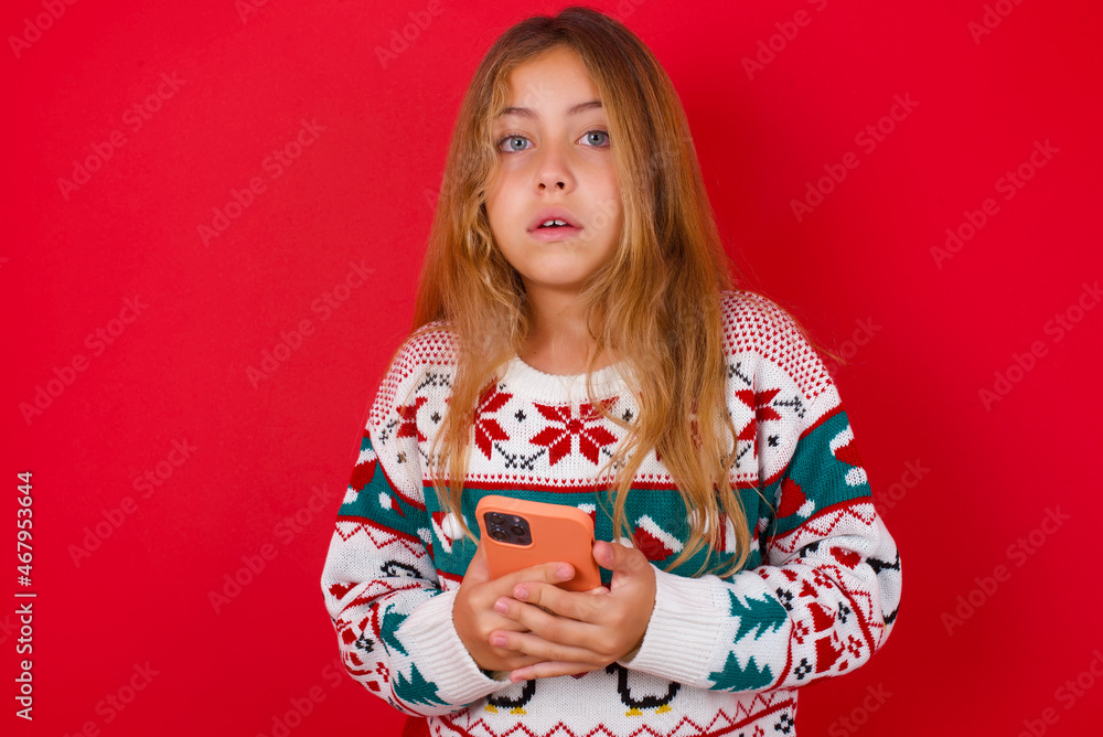 Photo of astonished crazy brunette kid girl in knitted sweater christmas over red background hold smartphone dislike feedback concept Photo of astonished crazy brunette kid girl in knitted sweater christmas over red background hold smartphone dislike feedback concept