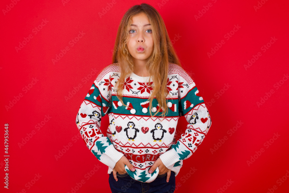 Portrait of lovely funny brunette kid girl in knitted sweater christmas over red background sending air kiss