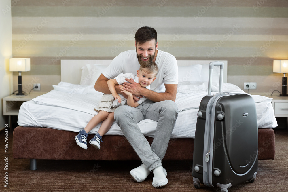 A young father and son sit on a bed in a hotel with a suitcase and laugh.