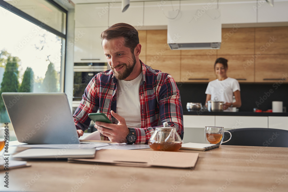 Happy smiling male in shirt looking to the screen of laptop while sitting at the wooden desk Happy smiling male in shirt looking to the screen of laptop while sitting at the wooden desk