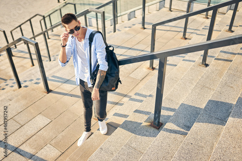 young man in sunglasses with a black backpack stands on the stairs leaning on the railing. © Ryzhkov Oleksandr