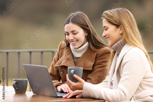 Two happy friends checking laptop in winter in a terrace