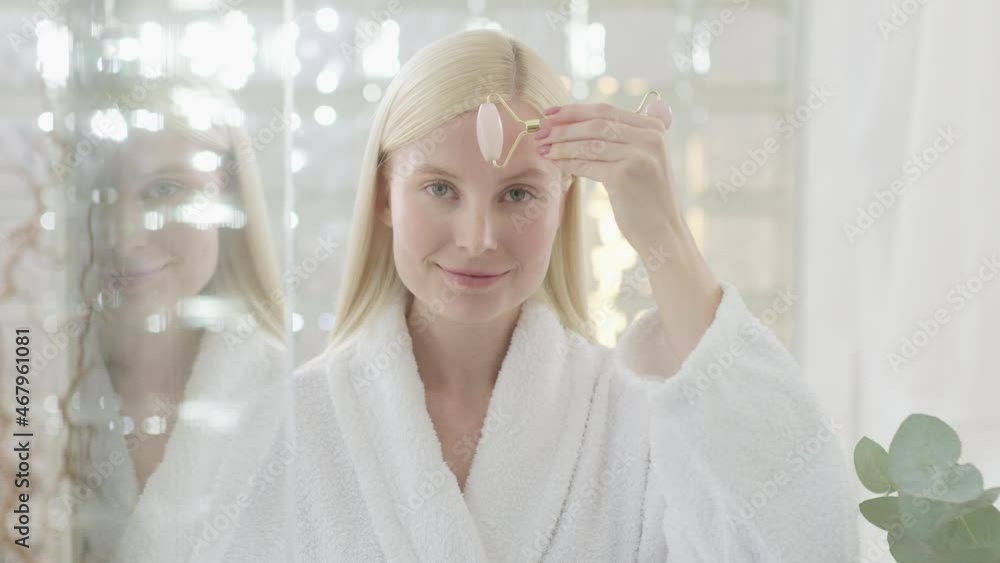 Portrait of Attractive Young Blond Woman Wearing Bathrobe With Natural Makeup With a Roller Massager Massages Her Face and Looking at Camera. Slow Motion Portrait of Attractive Young Blond Woman Wearing Bathrobe With Natural Makeup With a Roller Massager Massages Her Face and Looking at Camera. Slow Motion