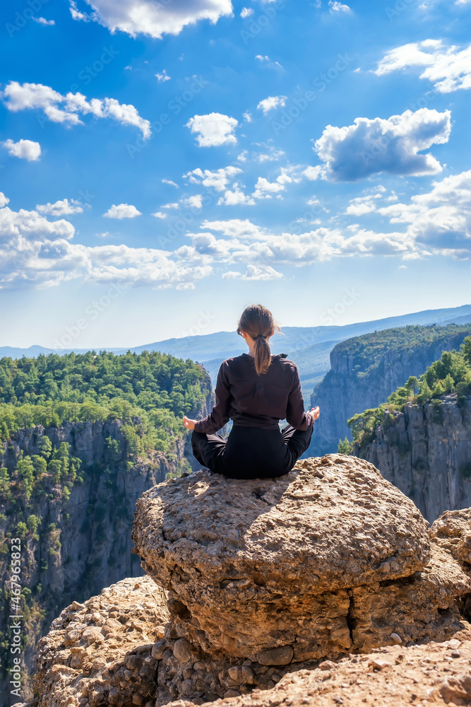 Back view of anonymous peaceful female sitting in lotus pose on edge of rocky cliff above mountains in highland during yoga session, vertical orientation, copy space