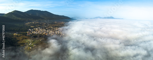 Morning fog over the valley, clouds from above, mountain village on the hillside, aerial view, drone shot.