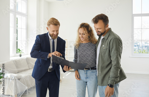 Happy married couple buying new house. Young husband and wife standing in white living room of partially furnished apartment and looking at contract that their real estate agent gives them to sign photo