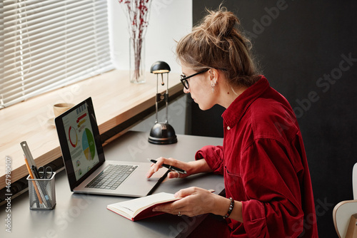 Fototapeta Young woman sitting at the table and using her laptop in her online work, she wo