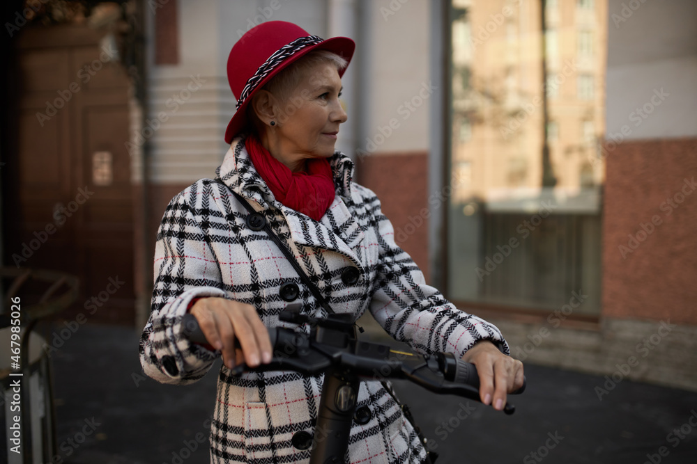 Elderly woman using e-scooter, standing against house with big windows in plaid trench coat and hat, looking aside, having stylish blonde short haircut. Active lifestyle. People and technology concept