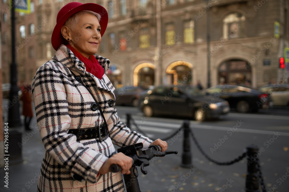 People and technology. Mature charismatic and attractive beautiful female in stylish clothes, red hat riding e-scooter enjoying city scape, central streets, architecture and big city life