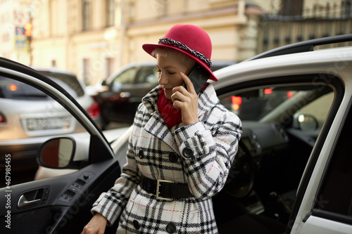 Attractive, pretty and stylish mature businesswoman getting out from car while talking on phone with her subordinates, giving the orders, wearing fashionable red hat. City lifestyle, busy days