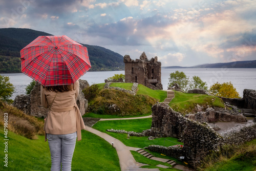 A tourist woman with a scottisch pattern umbrella looks at the famous Urquhart Castle at Loch Ness during autumn time, Scotland photo