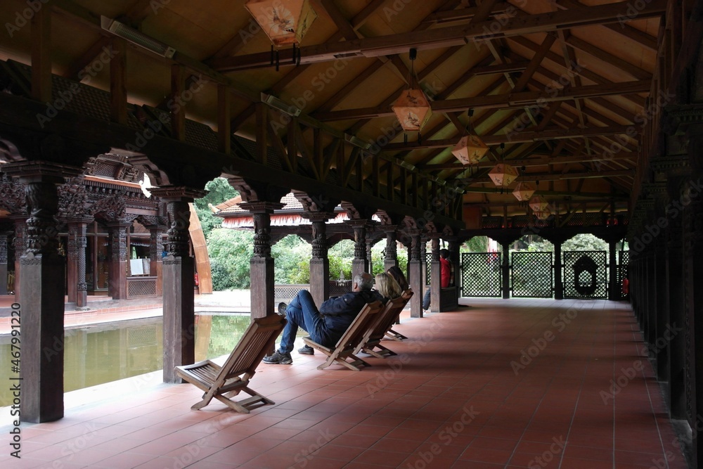 Fototapeta premium place of calmness and meditation: сourtyard of the tibetan temple with water pool, resting sitting people, wooden chairs in the Nepal Himalaya Park Stiftung in Bavaria, Germany.