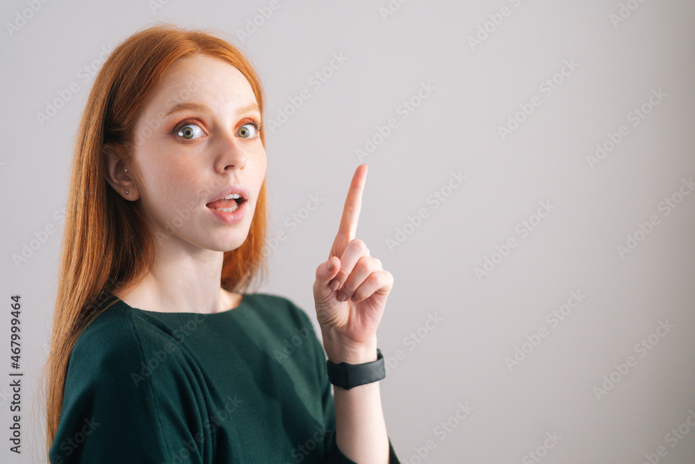 Portrait of amazed redhead young woman model pointing up with finger, looking at camera on white background. Medium shot of red-haired lady with natural beauty posing in professional studio. Portrait of amazed redhead young woman model pointing up with finger, looking at camera on white background. Medium shot of red-haired lady with natural beauty posing in professional studio.