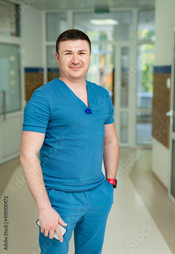 Young man wearing blue uniform posing for camera in modern hospital. Handsome medical specialist in uniform.