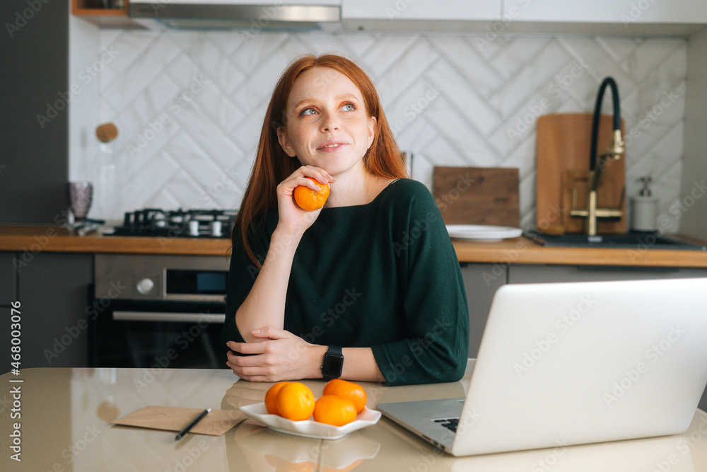 Medium shot portrait of happy smiling young woman sitting at table with laptop and holding tangerine in hand, dreaming looking up. Positive freelancer female working online from home office.