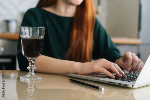 Close-up cropped shot of female freelancer working typing on laptop computer sitting at table with glass of alcoholic beverage in remote workplace. Young woman writing post comments in social media.