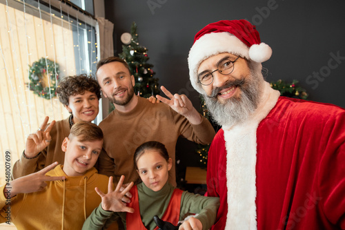 Selfie of smiling Santa Claus with gray beard posing with cheerful family showing peace signs while celebrating Christmas