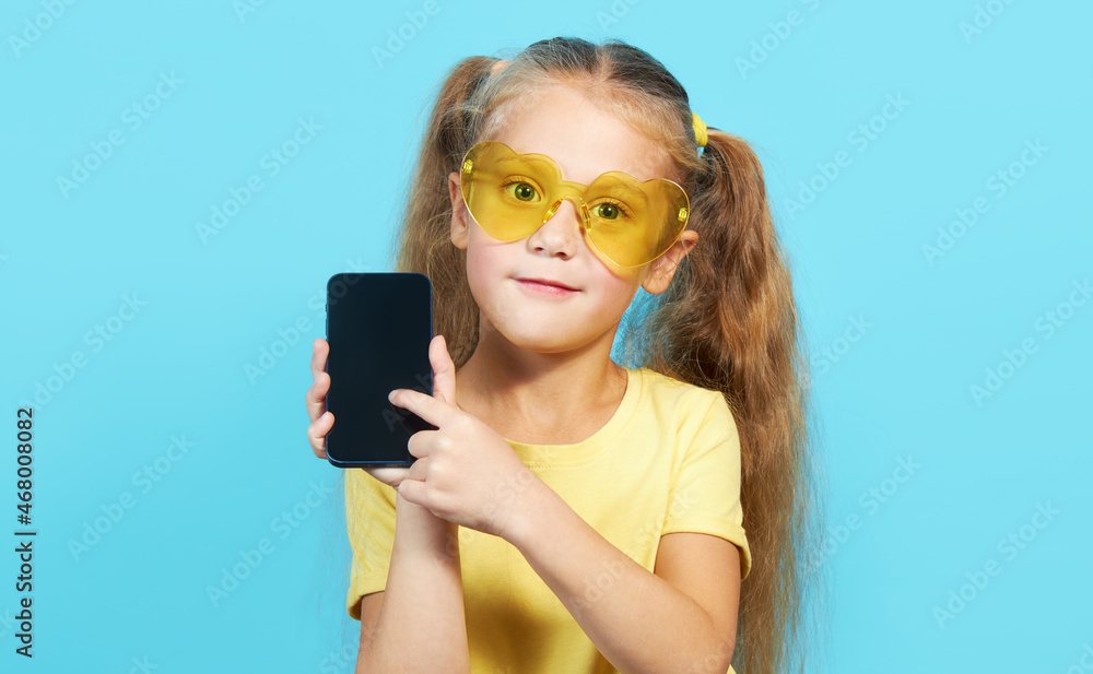 Awesome mobile app. A little surprised child girl in shape sunglasses pointing at cell phone with shocked face, online service, Parental control app. Studio shot blue background