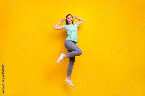 Full size photo of crazy brunette hairdo millennial lady jump v-sign wear t-shirt jeans sneakers isolated on yellow background