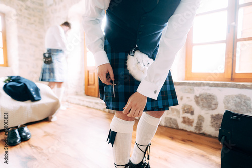 A man in Scottish national dress prepares for a wedding ceremony in a hotel room 