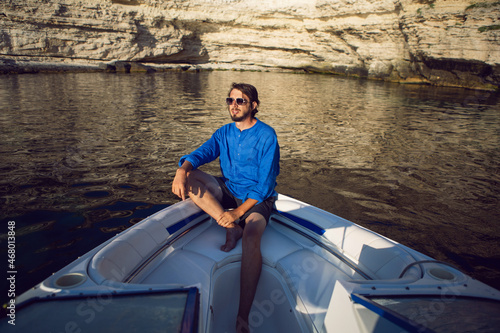 rich free man in sunglasses and a blue shirt sits on the bow of white boat